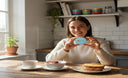 A smiling person in a cozy kitchen at breakfast time, holding a clear night guard case beside a cup of tea — bright, clean lighting, lifestyle feel